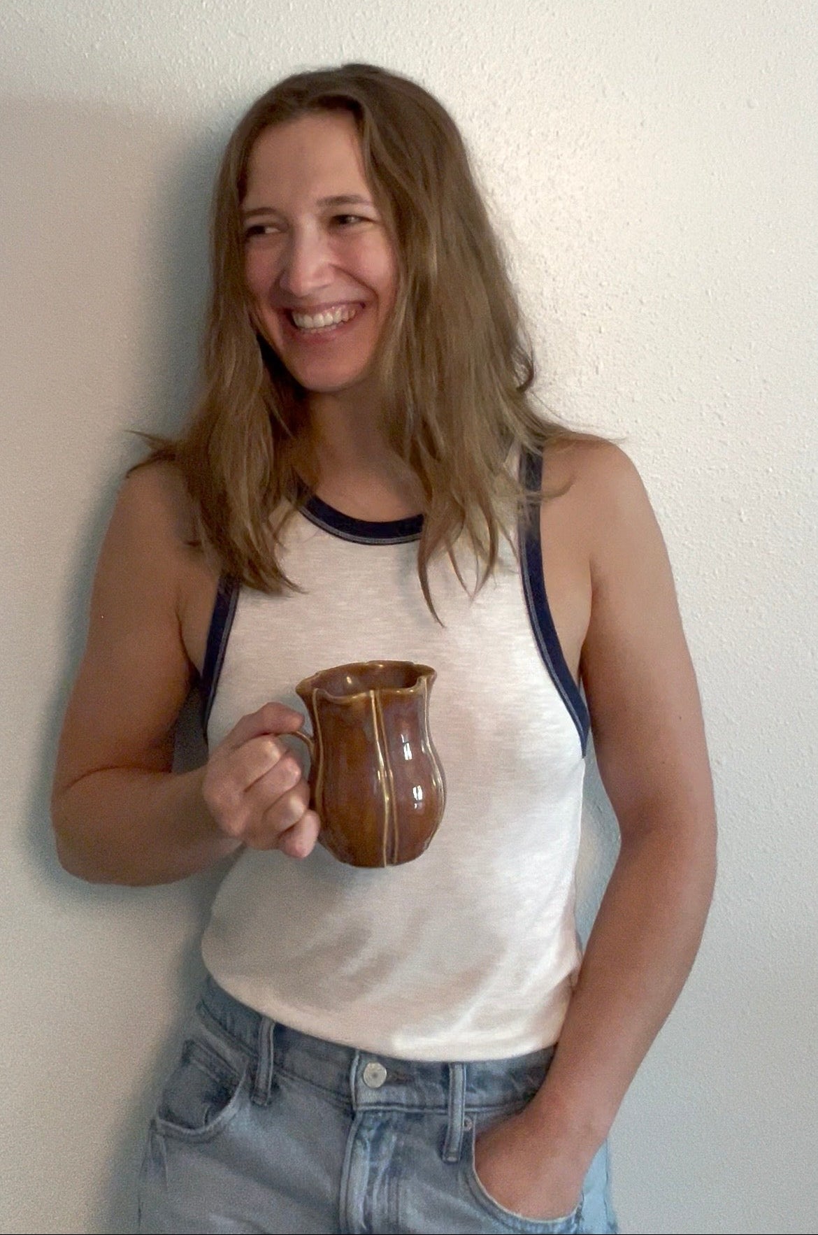 A woman with long light brown hair smiling while holding a brown ceramic mug, wearing a white tank top with dark navy trim and light blue jeans, standing against a plain off-white wall.